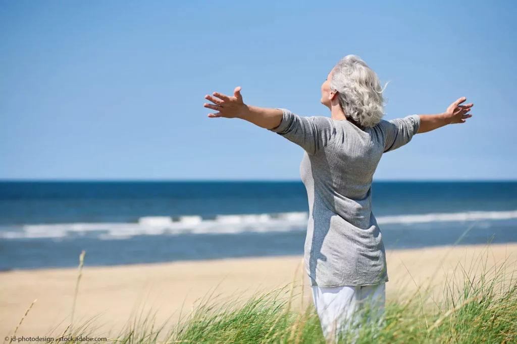 Praxis für Ergotherapie Boettcher in Greifswald - Behandlungsbereich -Big-Therapie-Bewegungstherapie-Ältere Frau mit grauem Haar steht am Meer -Blick Richtung Wasser und breitet die Arme aus